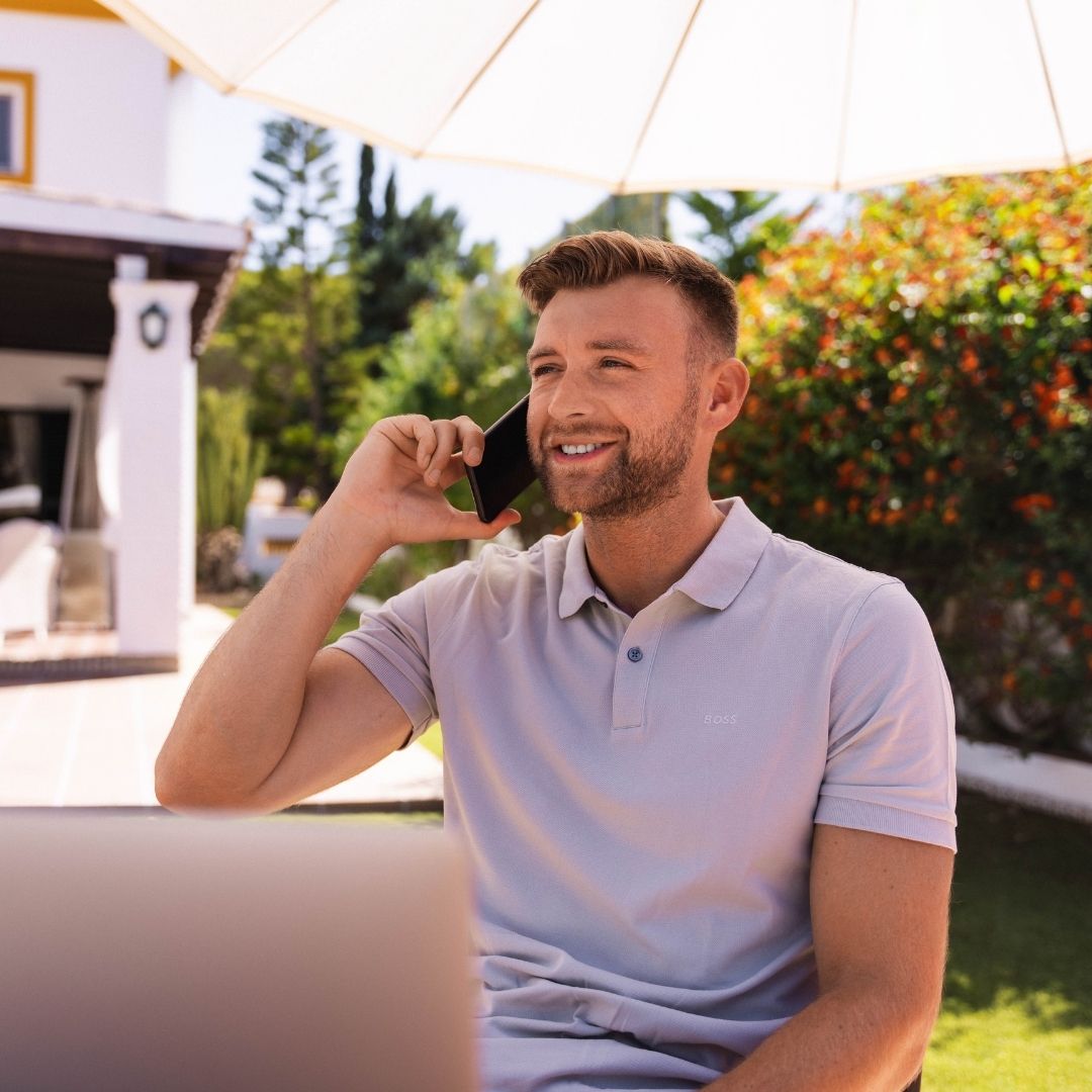 Junger Mann arbeitet am Laptop mit Telefon in der Hand im Garten bei gutem Wetter